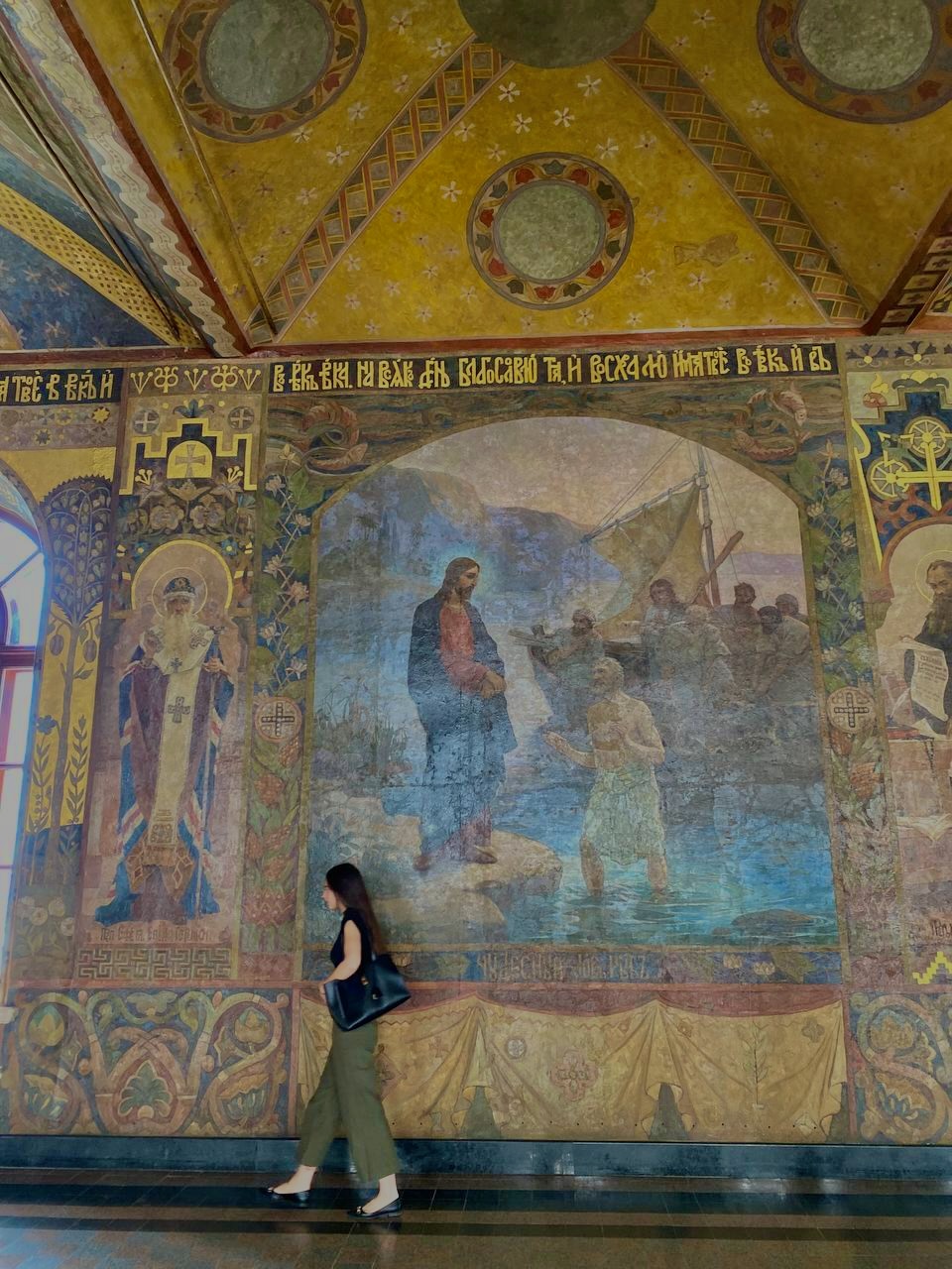 Sophia walking past a monumental Byzantine fresco inside an Orthodox cathedral