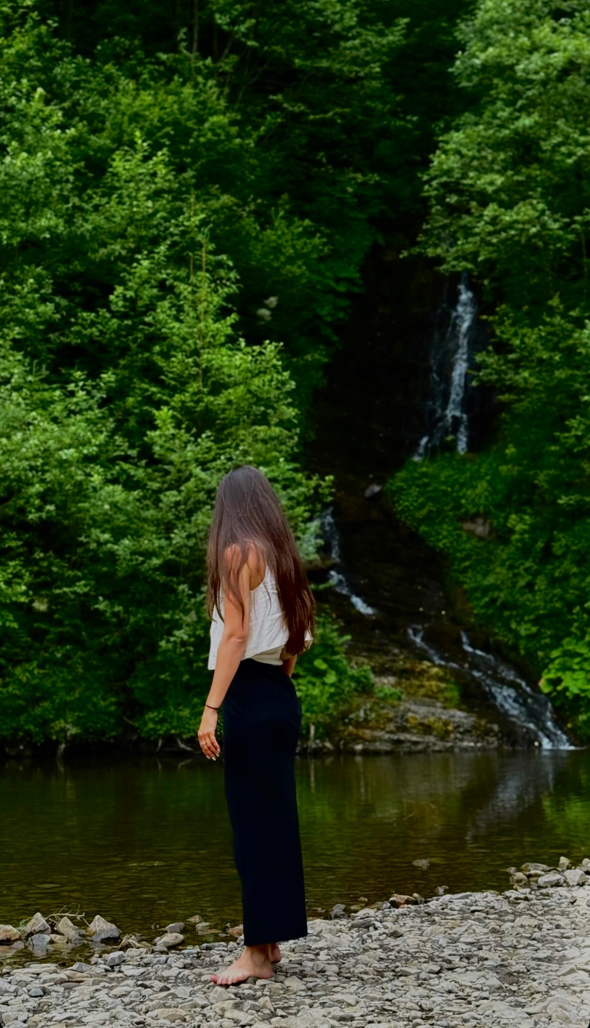 Sophia standing barefoot by a river with a waterfall in the Carpathian forest