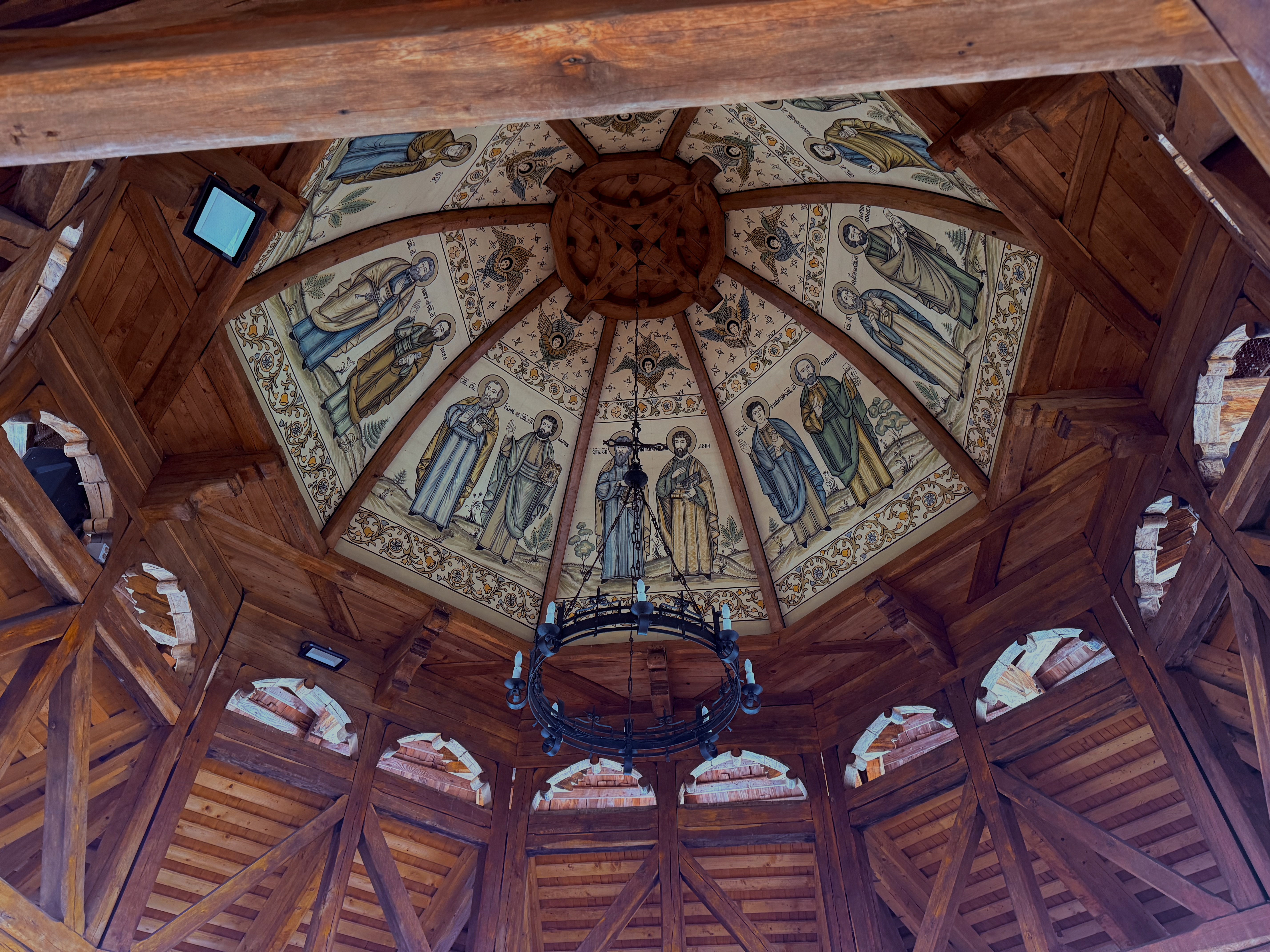 Painted dome ceiling inside a wooden monastery pavilion, saints depicted in warm gold tones around an iron chandelier