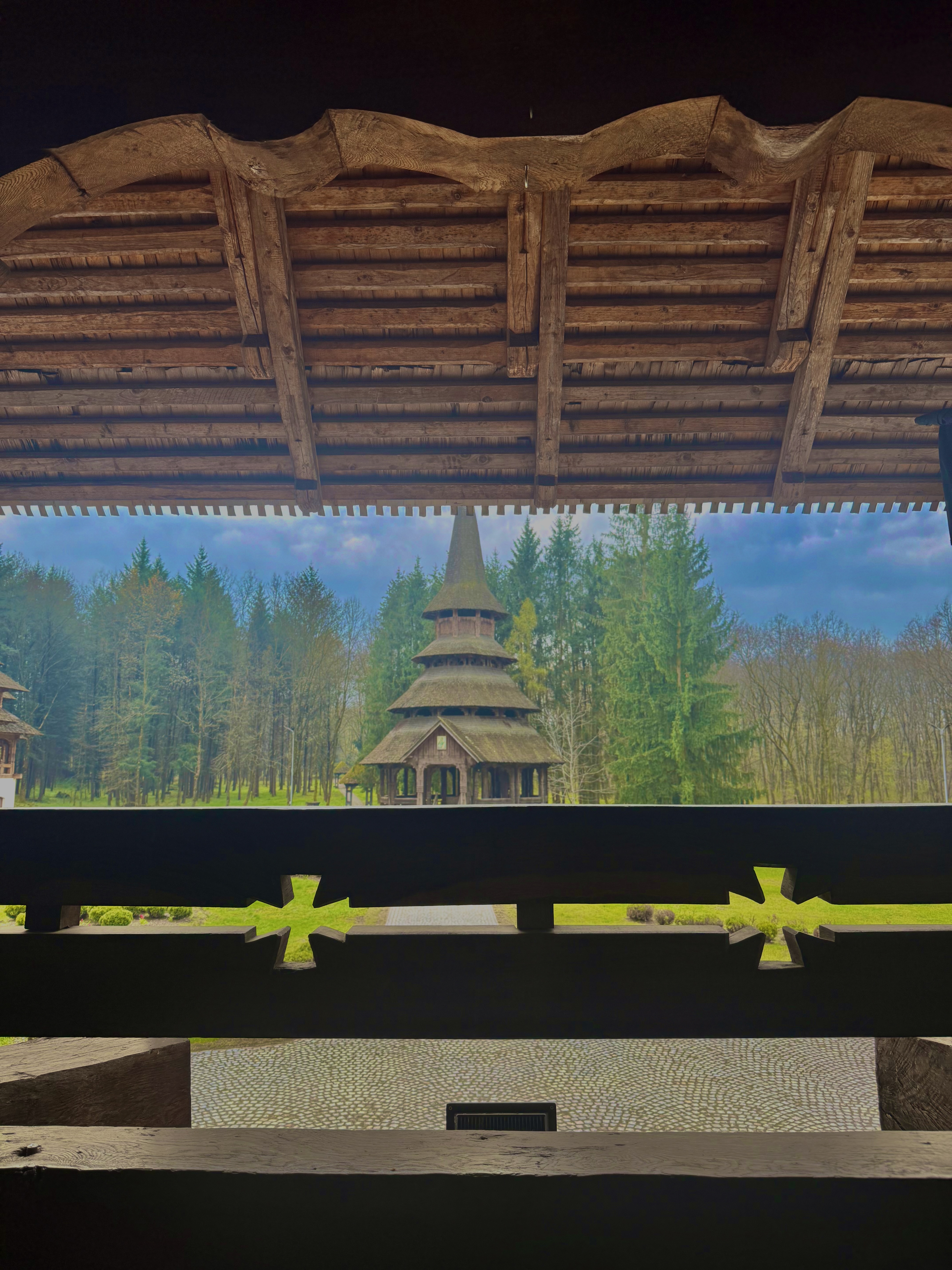 Wooden church spire framed through an ornate gate pavilion, spring-green forest in the distance