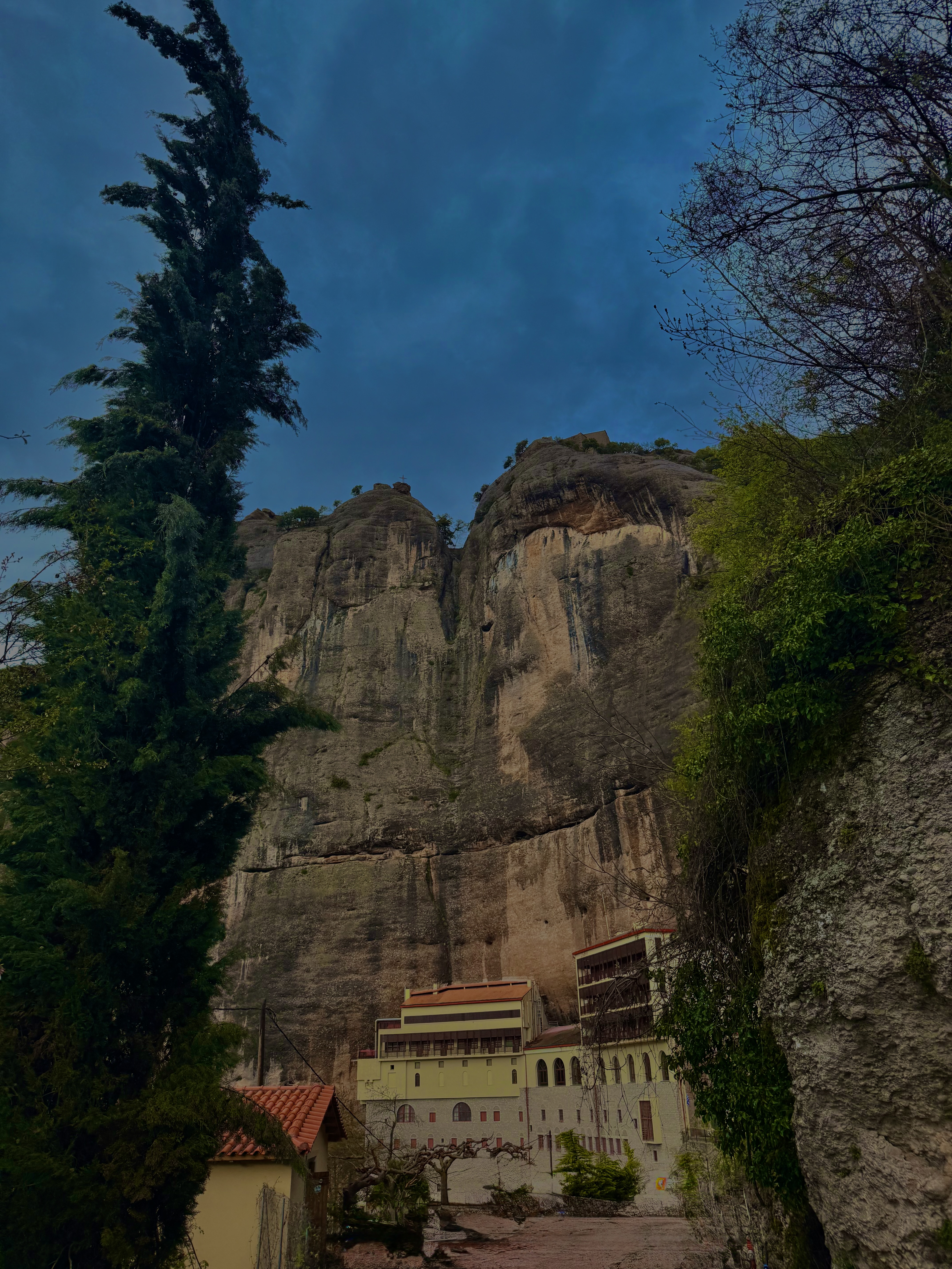 Monastery nestled at the base of towering limestone cliffs, cypress trees framing the ancient stone walls