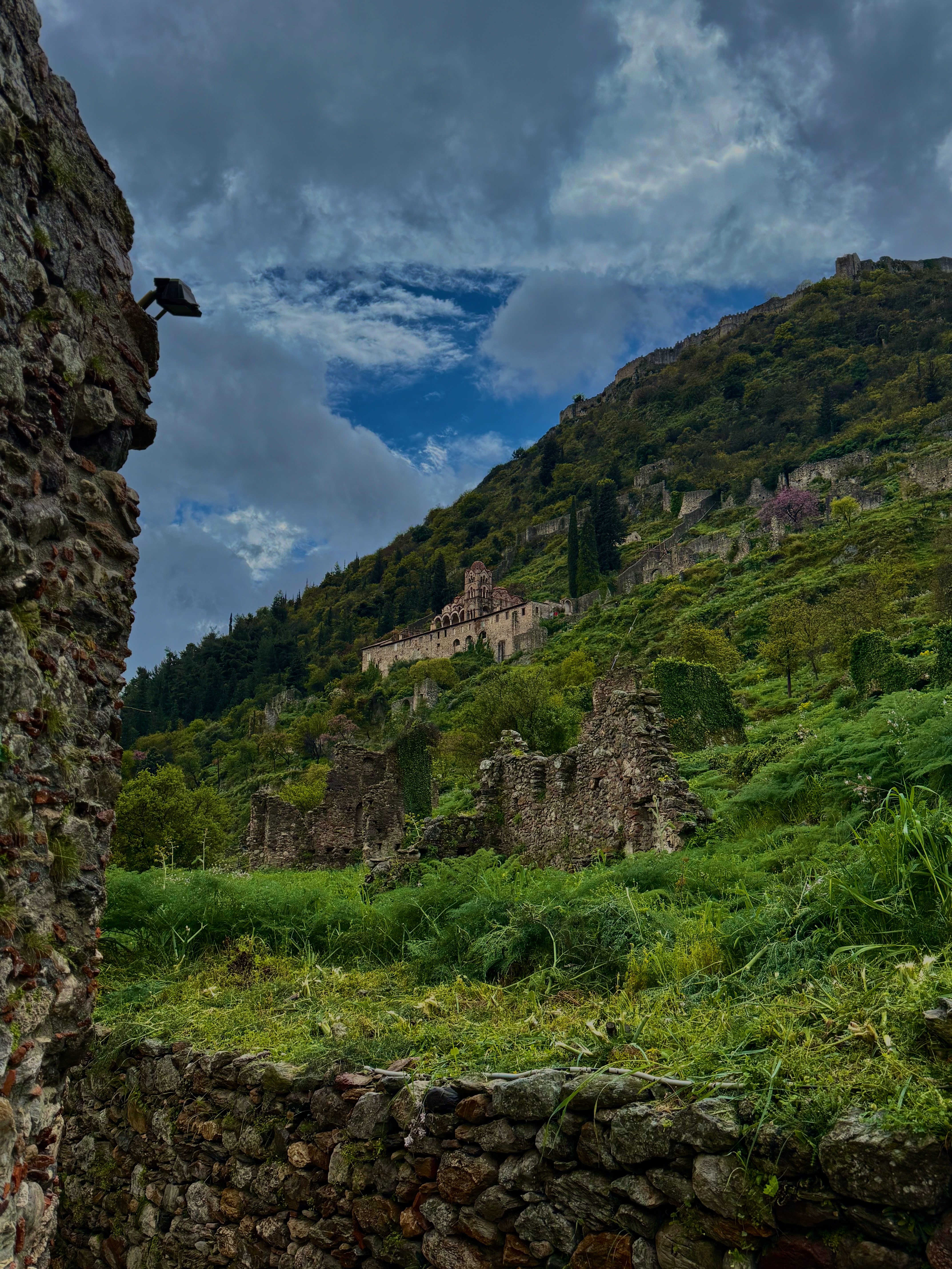 Byzantine ruins on a green hillside with a monastery perched above, stone walls covered in ivy